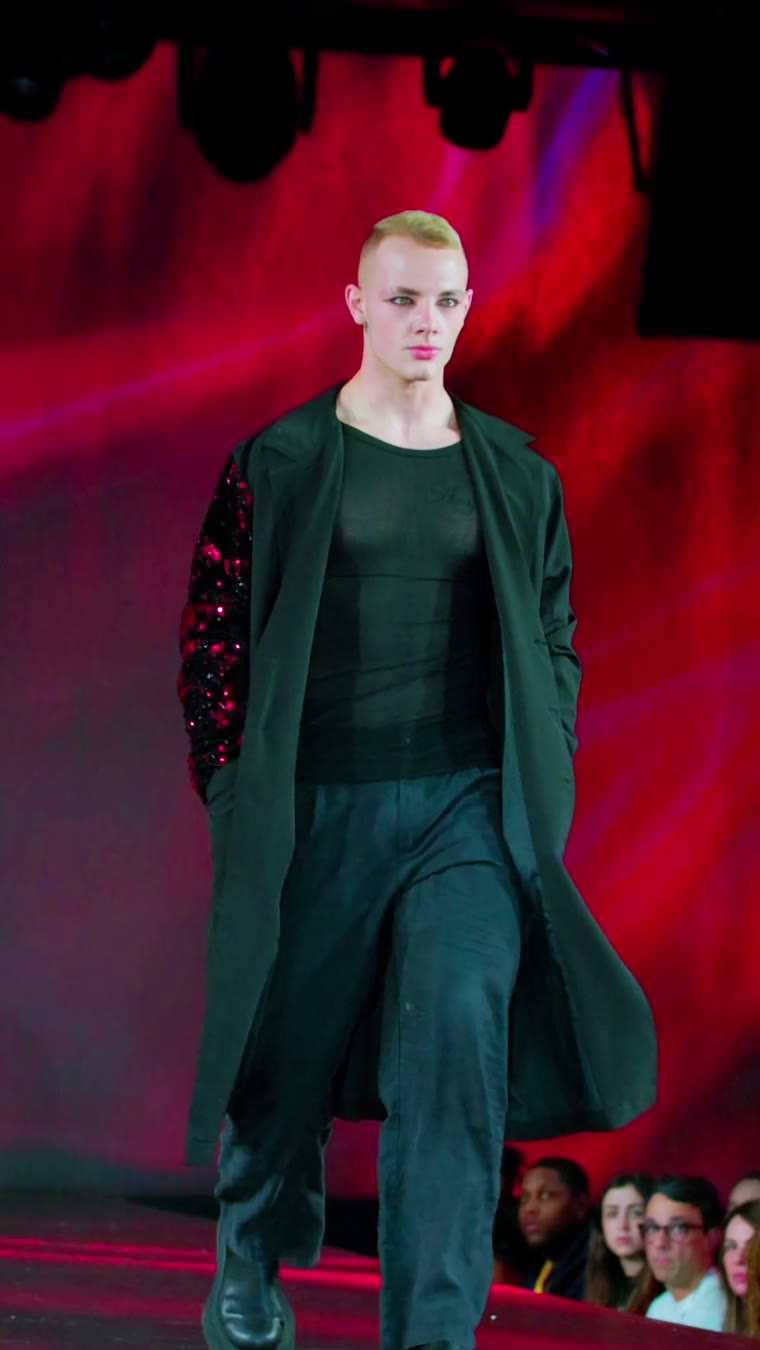 A male model in a long green coat on a red-lit runway
