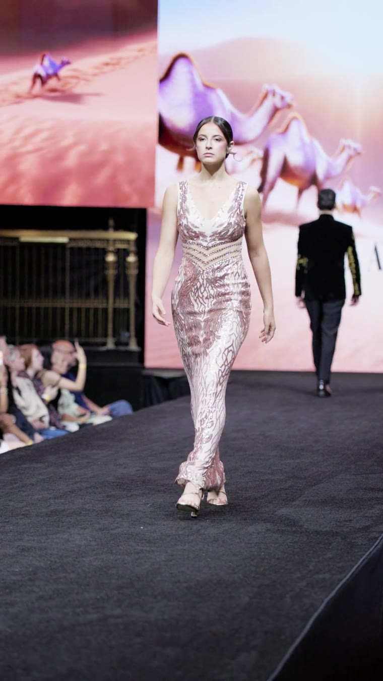 A model in a pink sequin gown on the runway, a desert scene on the screen behind her