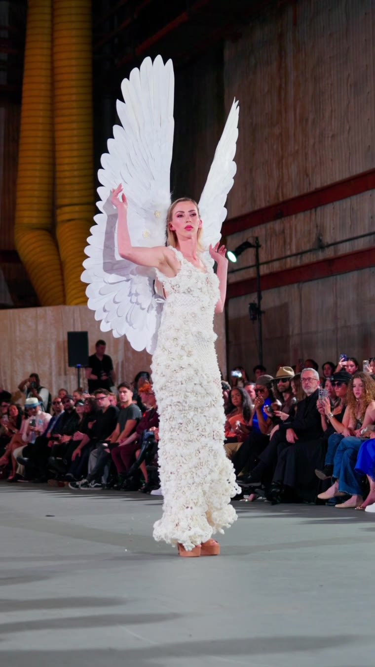A model in a white gown with big angel wings on the runway